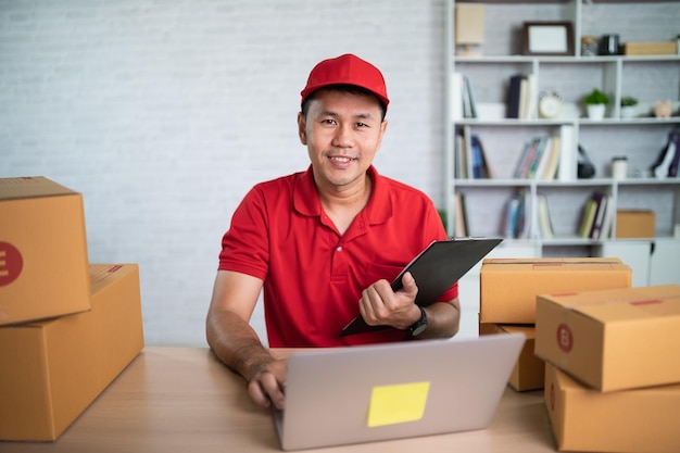 Foto aziatische bezorger werknemer glimlachend in rood uniform werk thuis controleren lijst pakketdozen voor het verzenden of vervoeren van pakketten per post levering transportconcept