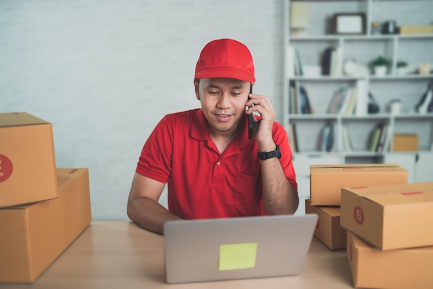 Aziatische bezorger werknemer glimlachend mobiele telefoon bellen naar klanten voor dozen voor het verzenden of vervoeren van pakketten per post Rood uniform werk thuis dragen