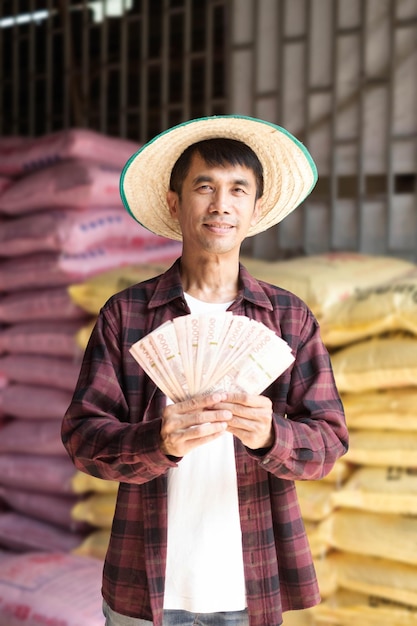 Aziatische boer met een rood shirt die een Thaise biljet met een zak chemische meststoffen vasthoudt in een magazijn