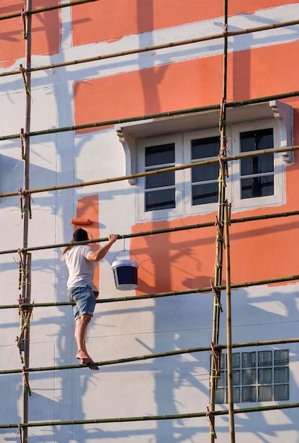 Aziatische bouwvakker op houten steiger schildert muur buiten het oude huisgebouw