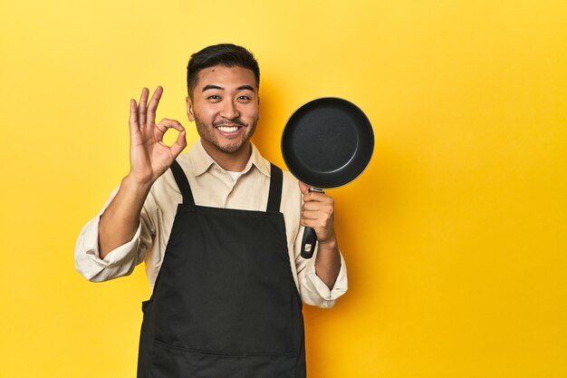 Aziatische chef-kok met een pan gele studio achtergrond vrolijk en zelfverzekerd met een ok gebaar