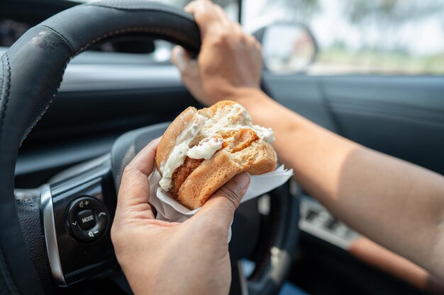 Aziatische dame die hamburger vasthoudt om in de auto te eten, gevaarlijk en riskeert een ongeluk