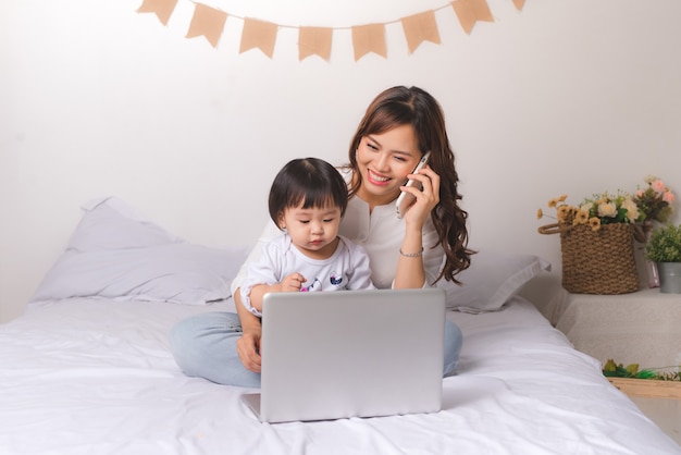 Aziatische dame in klassiek pak praat op de mobiele telefoon en werkt thuis op een laptop met haar babymeisje.