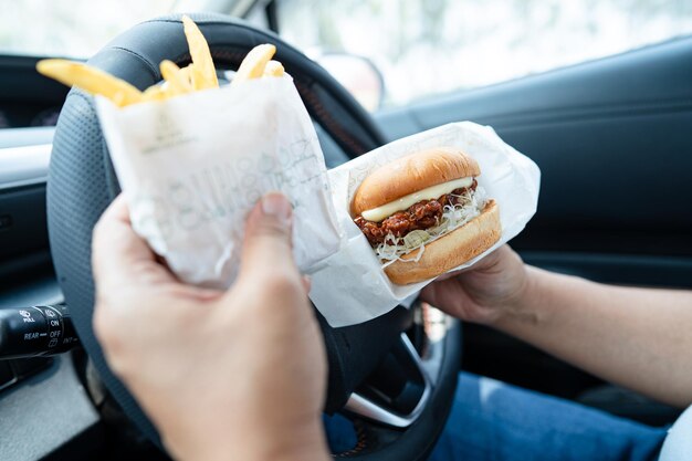 Aziatische dame met hamburger en friet om in de auto te eten gevaarlijk en risico op een ongeluk