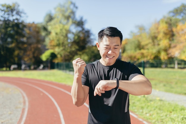 Aziatische die in de zonnige dag van het stadion loopt, kijkt naar de smartwatch en is tevreden met het resultaat