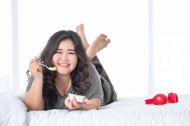 Aziatische dikke vrouw die van het eten van oranje cake op bed geniet