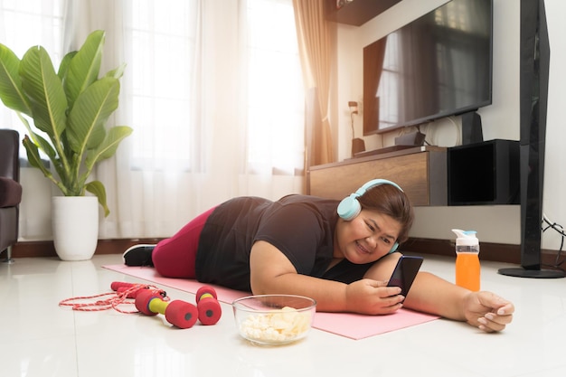 Aziatische dikke vrouw geniet van het eten van junkfood en ontspant zich door muziek van hoofdtelefoon en smartphone, concept voor sport en recreatie.
