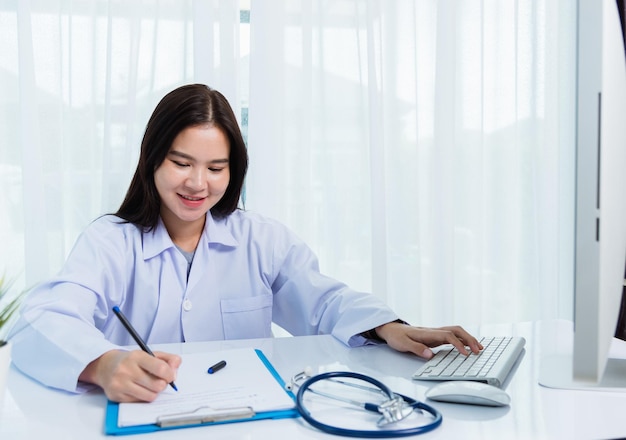 Aziatische dokter jonge mooie vrouw die lacht met behulp van werken met pc-desktopcomputer en haar schrijven iets op papierwerk of klembord wit papier op ziekenhuis bureau kantoor, gezondheidszorg medisch concept