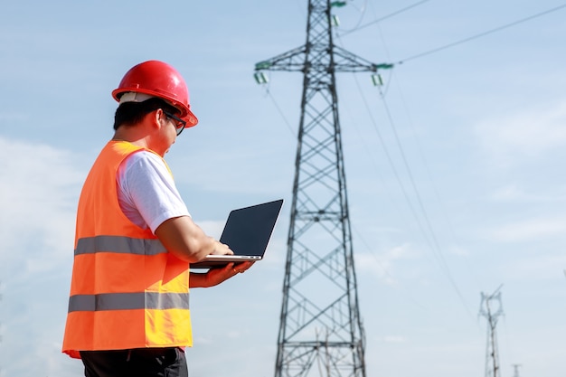 Aziatische elektrische ingenieursarbeider in standaard veiligheidsuniform met behulp van een mobiel die bij een elektriciteitscentrale staat om de hoogspanningspaal voor elektriciteit te inspecteren.