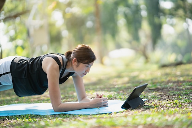 Aziatische fitness vrouw doet een yoga volgens een videoclip. Aziatische vrouw die tablet gebruikt en yoga in park doet