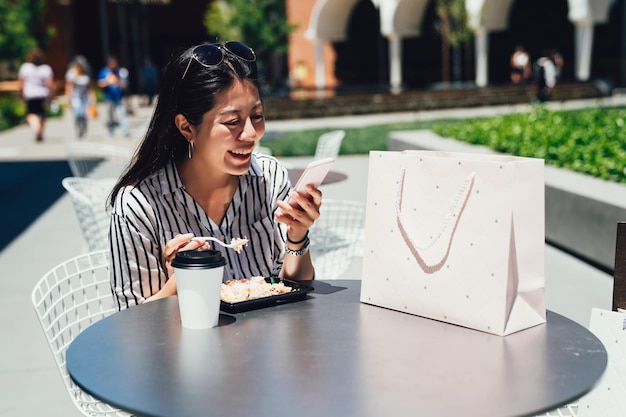 Aziatische gelukkige jonge vrouw die voedsel in Amerikaanse stijl eet in openluchtcafé en een bericht leest op een smartphone die vrolijk glimlacht. meisje aan het lunchen met koffiezakken in winkelcentrum stanford.