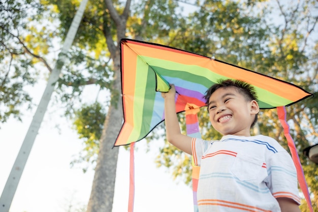 Aziatische gelukkige kinderenjongen met een vlieger die rent om door te vliegen in het park bij de zomerzonsondergang buitenshuis
