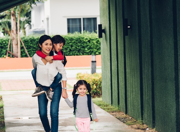 Aziatische gelukkige moeder loopt van huis weg terwijl ze haar kinderen hand in hand houdt en naar school gaat