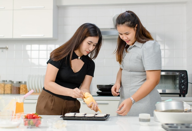 Aziatische gelukkige professionele vrouwelijke bakkerij bakkerij chef-kok huisvrouw met goede vrienden staan glimlachend helpen met het gebruik van buis zak knijpen slagroom versieren topping het bereiden van cupcakes in de keuken