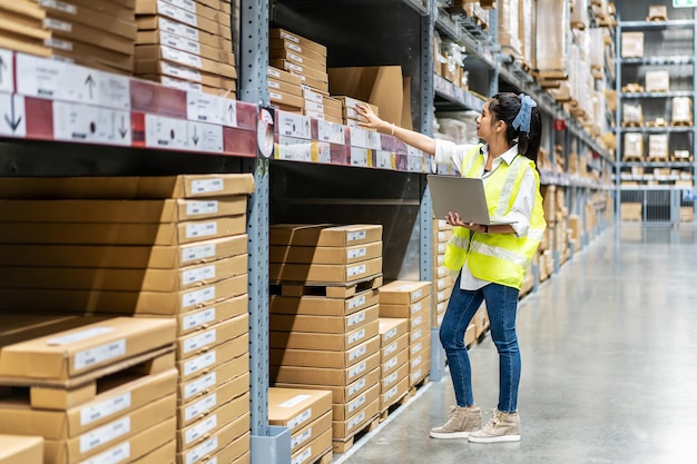 Aziatische ingenieursvrouw controleert voorraadgegevens op tabletcomputer voor het aanvinken van dozen met logistiek op planken met goederenachtergrond in magazijn, logistieke en zakelijke export