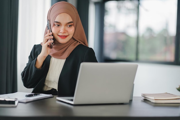 Aziatische islamitische zakenvrouw met hijab die telefoon gebruikt terwijl ze aan de computer werkt in het kantoor