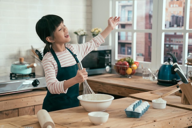 aziatische japanse kleine meid chef-kok draagt een schort dansen zingen geniet van muziek sluit de ogen en steekt de hand op terwijl hij zweepslagen door in de kom te kloppen. dochter bereidde het bakken van broodcake voor een gelukkige moederdag thuis