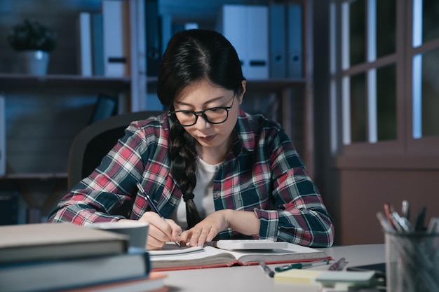 Aziatische Japanse vrouwelijke tienerstudent die boeken leest en 's avonds laat studeert op een donkere plaats in het appartement. leer- en onderwijsconcept. studente met een bril die om middernacht thuis huiswerk maakt