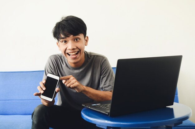Aziatische jeugd zit op de bank voor de laptop en wijst naar zijn mobiele telefoon