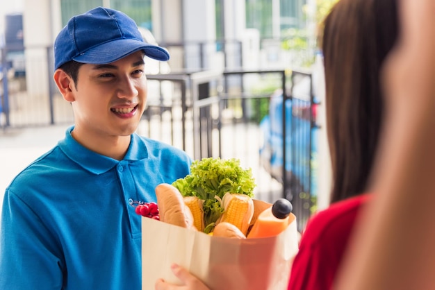 Aziatische jonge bezorger in uniform die boodschappen doet en vers voedsel in papieren zak geeft aan vrouwelijke klant die aan de huisdeur ontvangt onder pandemisch coronavirus, terug naar nieuw normaal concept