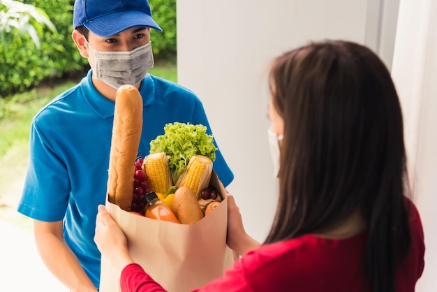 Aziatische jonge bezorger in uniform draagt beschermend gezichtsmasker hij doet boodschappen en geeft vers voedsel aan vrouwelijke klant die voorhuis ontvangt onder pandemisch coronavirus, terug naar nieuw normaal concept