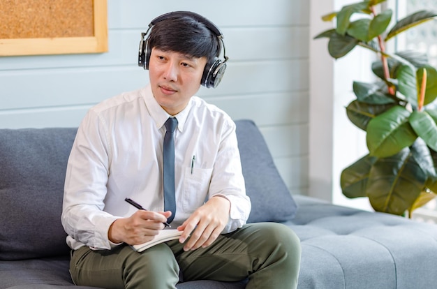 Aziatische jonge knappe professionele succesvolle mannelijke zakenman werknemer in formele zakelijke overhemd en stropdas zittend op een gezellige bank luisteren naar streaming muziek online surfen op internet met smartphone.