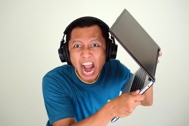 Aziatische jonge man met een koptelefoon met boze uitdrukking en gooit een laptop op wit wordt geïsoleerd
