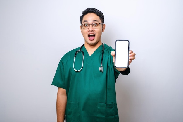Aziatische jonge man verpleegster arts met scrubs en stethoscoop kijkend naar camera met een leeg smartphonescherm die toepassing aanbeveelt
