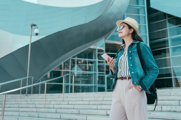 aziatische jonge meisjestoerist die de trap uit de concertzaal loopt in la die de online kaart-app van de mobiele telefoon controleert. mooie college vrouw student verlaten universiteit met rugzak chatten op mobiel.