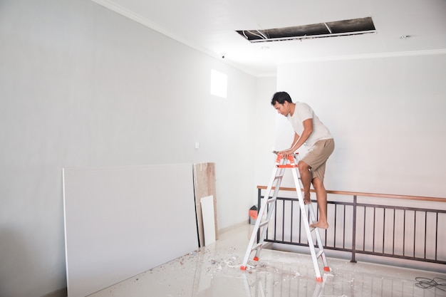 Aziatische jonge mens die een ladder gebruikt om het gebroken plafond thuis te herstellen