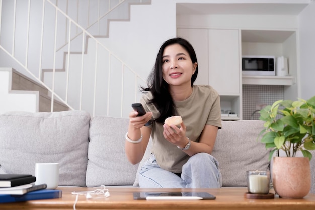 Aziatische jonge mooie vrouw kijken naar leuke film op televisie thuis Aantrekkelijk casual meisje voelt zich gelukkig en ontspannen zitten op de bank plezier kijken komedie video op TV in huis Activiteit levensstijlen concept