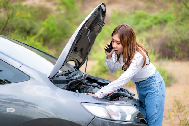 Aziatische jonge ongelukkige vrouw die op een celtelefoon voor de open kap opgesplitste auto bij de landweg telefoneren voor hulp spreekt.