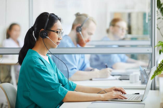 Aziatische jonge operator in uniform en hoofdtelefoon aan de tafel zitten en typen op laptop in het ziekenhuis