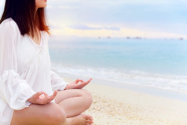 Aziatische jonge vrouw beoefent yoga op het strand in de buurt van de zee onder zonlicht bij zonsopgang, ontspanning voor gezondheid midden in de natuur met geluk en vrede, blanco voor de achtergrond