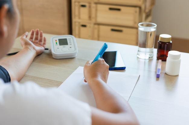 Aziatische jonge vrouw controleert zelf de bloeddruk en hartslag met een digitale manometer en registreert thuis op papier. gezondheid en medisch concept.