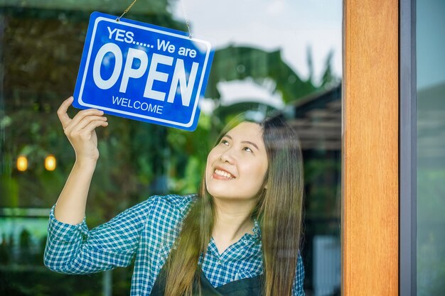 Aziatische jonge vrouw die een open bord bij de winkelglazen zet om de klant te verwelkomen