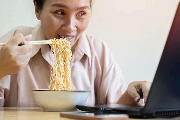 Foto aziatische jonge vrouw die instant noedels eet terwijl ze naar een video kijkt.