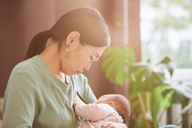 Aziatische jonge vrouw die naar haar babymeisje kijkt terwijl ze haar voedt in de slaapkamer