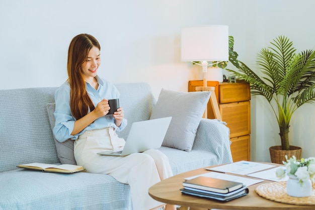 Aziatische jonge vrouw die thuis werkt met behulp van computer en koffie drinkt in haar kamer, documentfinanciering en online conferentievergadering voor nieuwe projecten