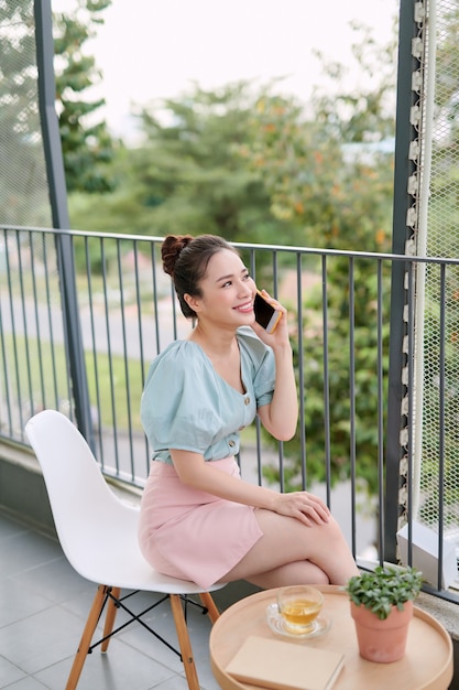 Aziatische jonge vrouw die van kop van koffie geniet tijdens het gebruik van telefoon op het balkon