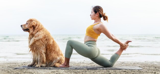 Aziatische jonge vrouw die yoga doet met haar hond op een yogamat op het strand Bannerformaat foto