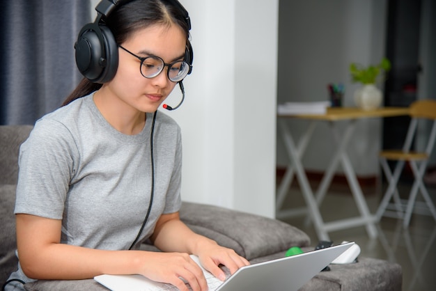 Aziatische jonge vrouw student met koptelefoon zittend op de bank kijken naar laptop gelukkig studeren online klasse college leren internet onderwijs, tienermeisje werk afstand op een laptopcomputer van huis