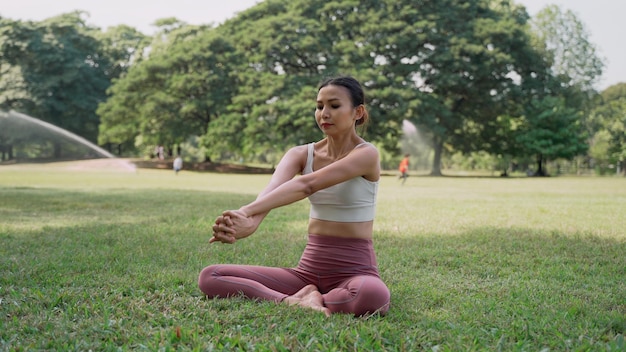 Aziatische jonge vrouw zittend op het gras in de lotuspositie en handen opsteken buiten in stadspark met de grote bomen achtergrond Achteraanzicht van vrouwelijke yoga beoefenen buiten op een zonnige dag