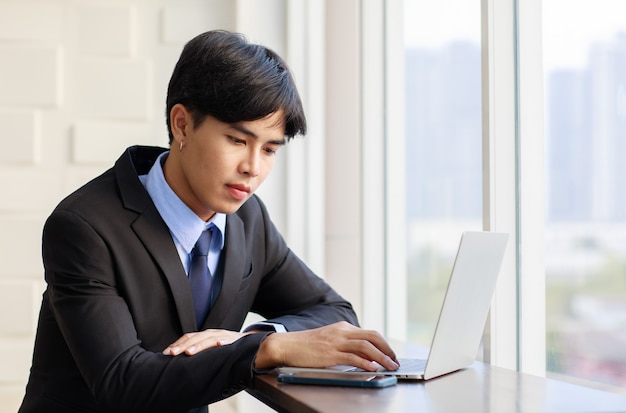Aziatische jonge zakenman is slim en knap in een zwart pak terwijl hij 's ochtends op het toetsenbord typt op een laptop in de buurt van het raam op kantoor.
