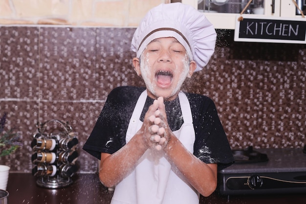 Aziatische jongen in chef-kokuniform lachend en klappend in de handen terwijl hij meel van deeg bestrooit in de keuken