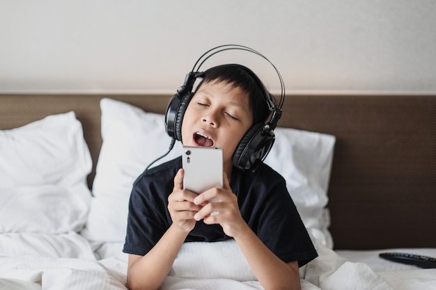 Aziatische jongen in hoofdtelefoon die muziek van smartphone luistert terwijl hij op het bed zingt