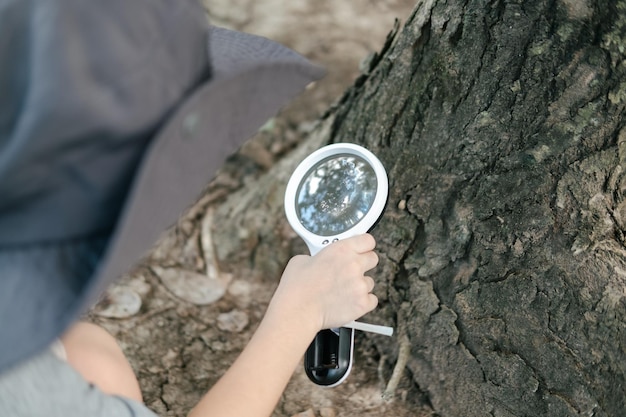 Foto aziatische jongen met een hoed in een bosverkennend pak gebruikt een vergrootglas om het boomgebied te onderzoeken