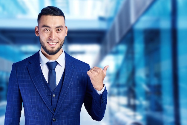 Aziatische Kazachse zakenman in pak lacht vriendelijk en toont vinger naar de zijkanten op kopieerruimte op een blauwe wazige achtergrond