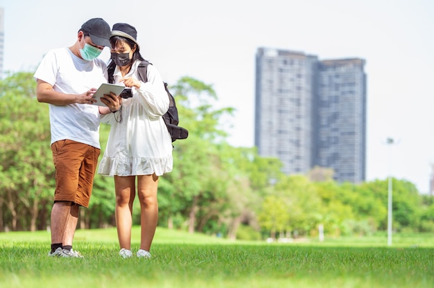 Aziatische koppels met gezichtsmasker op zoek naar toeristische informatie via tablets met geweldige plaatsen om te bezoeken in de stad met bouw- en parkachtergrond. Technologie en reizen concept. Nieuw normaal thema