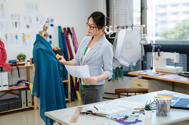 Aziatische Koreaanse vrouw kleermaker in naaister workshop werken. stijlvolle vrouwelijke modeontwerper met meetlint op nek kijken naar mannequin en schetspapier in studio. meisje personeel controleren met loting